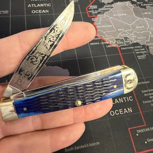 May include: A pocket knife with a silver blade featuring an engraved design and a blue handle. The handle has a textured appearance. The knife is open, revealing the blade's intricate details. A horseshoe emblem is visible on the handle.