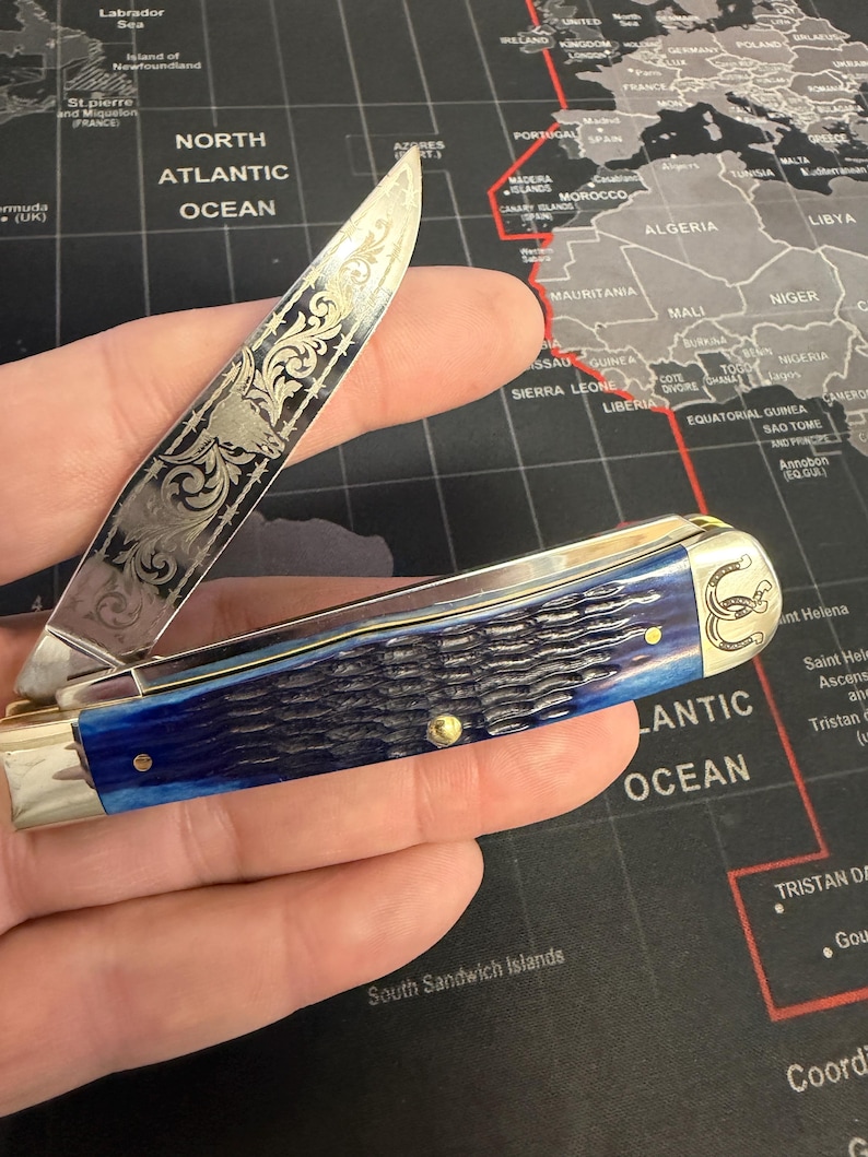 May include: A pocket knife with a silver blade featuring an engraved design and a blue handle. The handle has a textured appearance. The knife is open, revealing the blade's intricate details. A horseshoe emblem is visible on the handle.