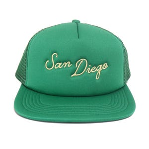 May include: Green trucker hat with the embroidered text "San Diego" in a cursive font. The hat features a green mesh back and a curved brim. The hat is a solid green color.