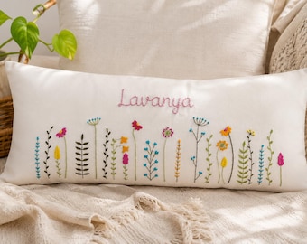 May include: A rectangular white throw pillow featuring hand-embroidered colorful wildflowers along the bottom edge and the name Lavanya stitched in pink thread across the center. The floral design includes various stems, leaves, and blossoms.