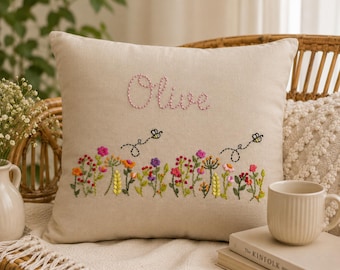 May include: A square beige throw pillow featuring hand-embroidered floral designs in pink, yellow, purple, and green. The name Olive is stitched in pink thread at the top, with two small black bees flying above the colorful wildflower garden scene.
