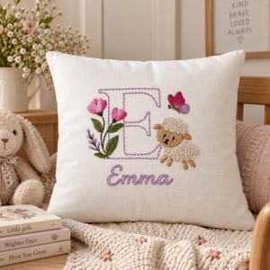 Può includere: Un cuscino quadrato bianco con una lettera E ricamata in viola, una piccola pecora, una farfalla rosa e fiori rosa. Il nome Emma è ricamato in corsivo viola sotto la lettera. Il tessuto presenta una trama intrecciata visibile.