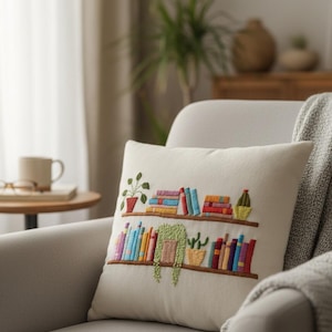 May include: Cream-colored throw pillow with embroidered bookshelves. The design features colorful books, potted plants, and a hanging plant. The pillow is placed on a gray armchair, with a blanket draped over the arm.
