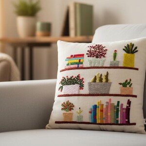 May include: Cream-colored decorative pillow with embroidered bookshelves. The shelves display colorful books and potted plants, including cacti and flowering plants. The pillow is on a gray sofa.