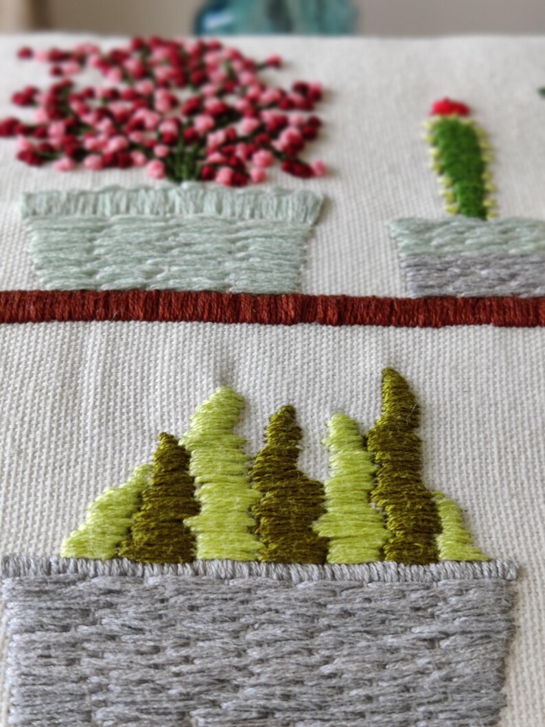 May include: Embroidered artwork featuring a botanical theme. The design includes a row of potted plants with green and red accents, and a row of stylized green trees. The artwork is on a cream-colored fabric.
