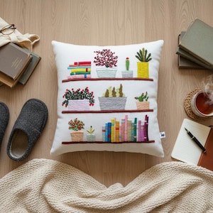 May include: A white decorative pillow with embroidered bookshelves, plants, and books. The image also includes a tote bag with glasses and books, gray slippers, a blanket, a cup of tea, and a notebook.
