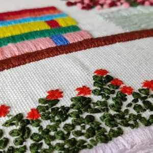 May include: Close-up of an embroidered textile featuring colorful stitched elements. The design includes a row of red flowers with green leaves, a series of stacked, colorful rectangles, and a brown border. The background is white.