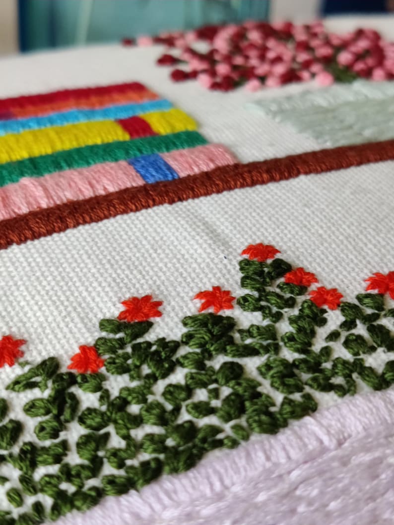 May include: Close-up of an embroidered textile featuring colorful stitched elements. The design includes a row of red flowers with green leaves, a series of stacked, colorful rectangles, and a brown border. The background is white.