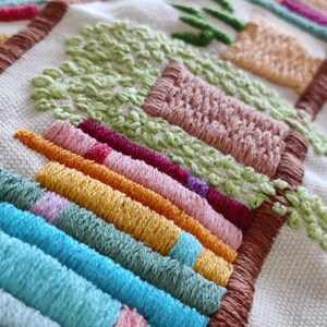 May include: Close-up of a colorful embroidered textile. The design features rows of vibrant, textured stitches in shades of blue, green, yellow, pink, and red. The embroidery includes what appears to be a landscape scene with trees and buildings.