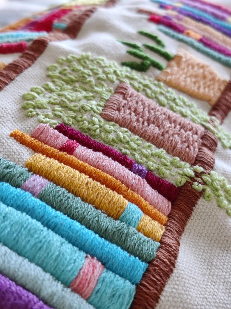 May include: Close-up of a colorful embroidered textile. The design features rows of vibrant, textured stitches in shades of blue, green, yellow, pink, and red. The embroidery includes what appears to be a landscape scene with trees and buildings.