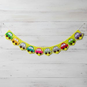 May include: A yellow felt banner with a string, decorated with colorful car shapes. The cars are in various colors, including green, orange, gray, blue, pink, and purple. The banner is designed for decoration.