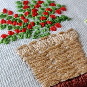 May include: Close-up of an embroidered design featuring a potted plant with red berries and green leaves. The pot is tan, and the embroidery is on a white fabric background. The design is framed with a brown border.