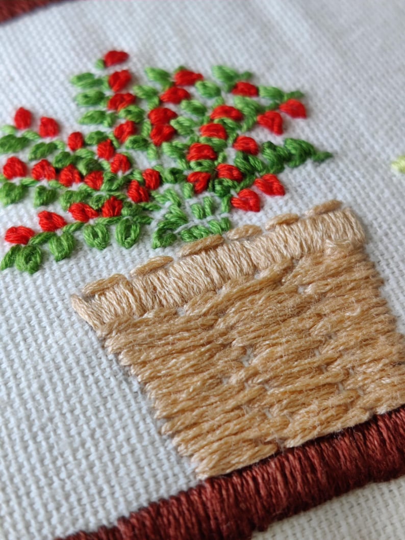 May include: Close-up of an embroidered design featuring a potted plant with red berries and green leaves. The pot is tan, and the embroidery is on a white fabric background. The design is framed with a brown border.