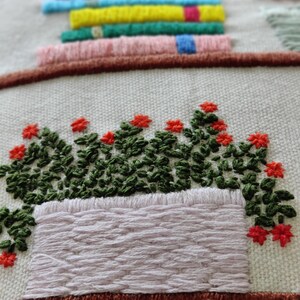 May include: Embroidered textile art featuring a stack of colorful books above a planter box filled with green foliage and red flowers. The embroidery uses various colors and textures, creating a detailed and vibrant design on a neutral background.