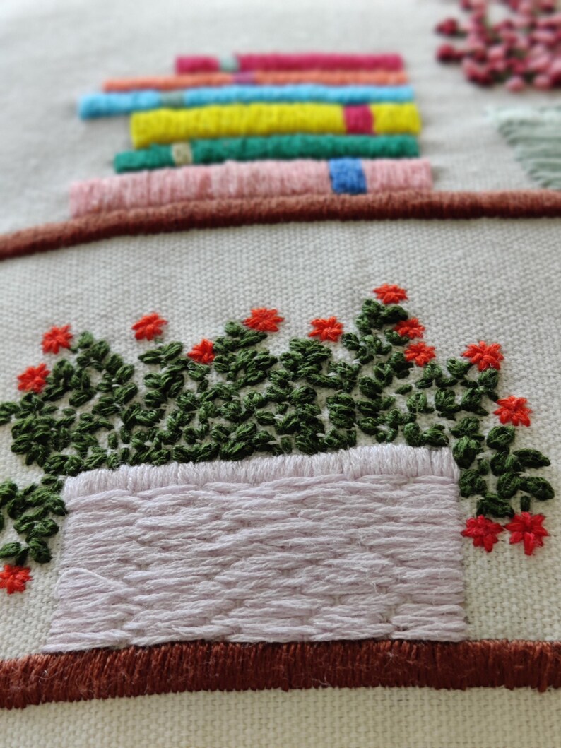 May include: Embroidered textile art featuring a stack of colorful books above a planter box filled with green foliage and red flowers. The embroidery uses various colors and textures, creating a detailed and vibrant design on a neutral background.