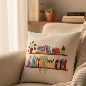 May include: Cream-colored throw pillow with an embroidered design of a bookshelf filled with colorful books and potted plants. The pillow is on a beige sofa, with a blanket draped over the arm.
