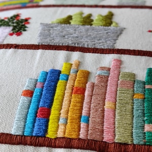 May include: Embroidered textile featuring a bookshelf design with colorful book spines. The books are in shades of blue, yellow, orange, pink, and green. Above the books are embroidered potted plants with red, green, and pink flowers.
