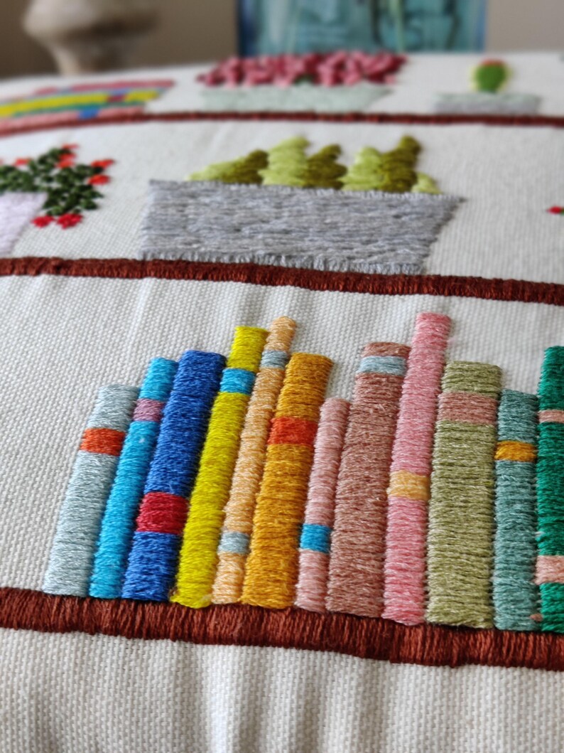 May include: Embroidered textile featuring a bookshelf design with colorful book spines. The books are in shades of blue, yellow, orange, pink, and green. Above the books are embroidered potted plants with red, green, and pink flowers.