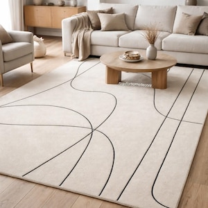 Puede incluir: Alfombra beige con diseño minimalista. Líneas negras curvas crean un patrón abstracto. La alfombra está sobre un suelo de madera en un salón.