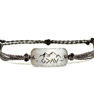 May include: A gray rope bracelet with a silver rectangular charm featuring a mountain range and the text "God is greater than the highs and lows".