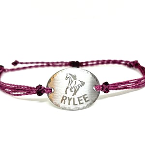 May include: A bracelet featuring a silver-toned oval metal charm with a hammered texture. The charm displays an engraved graphic of a horse above the name RYLEE in capital letters. The charm is attached to a dark magenta cord with adjustable knots.