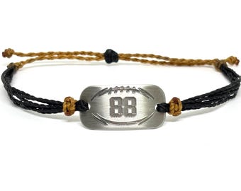 May include: A bracelet featuring a brushed silver-tone rectangular metal plate engraved with the number 88 and a football graphic. The plate is attached to black cord strands secured by brown knotted accents, with a brown cord adjustable closure.