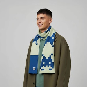 May include: A light green and blue scarf with a pixelated design. The scarf features a repeating pattern of blue pixelated figures against a light green background. The scarf is made of a soft material and is worn around the neck.