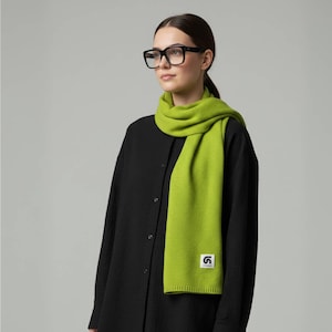 Può includere: Una donna indossa una sciarpa verde brillante e occhiali neri. La sciarpa è di colore pieno e ha una piccola etichetta bianca con un logo nero in basso. Indossa una camicia nera abbottonata.