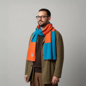May include: A color block scarf in orange and turquoise blue. The scarf has a small square logo on one end. The person in the image is wearing a brown sweater and a green coat.