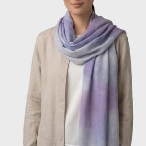 Puede incluir: Una mujer lleva una bufanda degradada lila y azul. La bufanda está drapeada alrededor del cuello y los hombros. La mujer lleva una camisa beige abotonada sobre una camiseta blanca y pantalones azules. La bufanda tiene un pequeño logotipo en la parte inferior.
