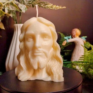 May include: A cream-colored candle shaped like a bust of a man with long hair and a beard. The candle is on a dark surface, with a white vase and greenery in the background. A small figurine is also visible.