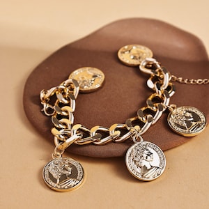 May include: Gold-toned bracelet featuring a chunky chain design with dangling coin charms. The coins display a profile image. The bracelet is displayed on a brown surface, showcasing the jewelry's details and shine.