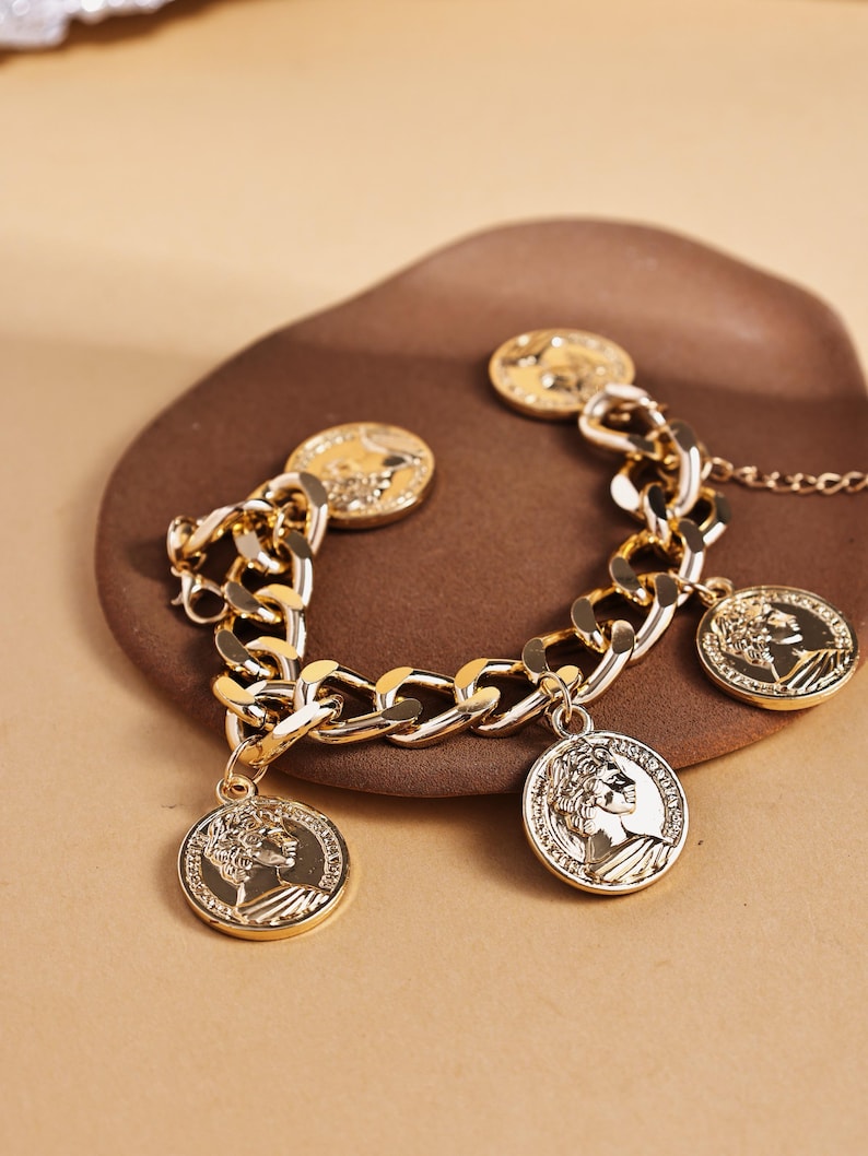 May include: Gold-toned bracelet featuring a chunky chain design with dangling coin charms. The coins display a profile image. The bracelet is displayed on a brown surface, showcasing the jewelry's details and shine.