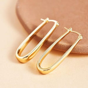 Könnte beinhalten: Zwei goldfarbene ovale Creolen. Die Ohrringe haben eine glatte, polierte Oberfläche und ein schlichtes, elegantes Design. Die Reifen sind etwa 3,8 cm lang und werden auf einem neutralen Hintergrund präsentiert.