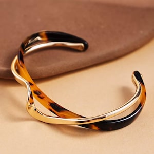 Peut inclure: Un bracelet manchette dor&eacute; avec un design tourbillonnant. Le bracelet pr&eacute;sente un motif &eacute;caille de tortue dans les tons marron et noir, avec une bande dor&eacute;e polie. Le design ouvert facilite le port.