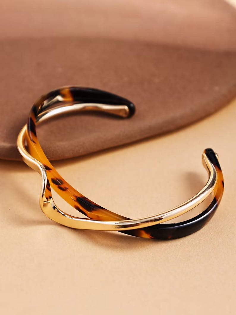 Peut inclure: Un bracelet manchette dor&eacute; avec un design tourbillonnant. Le bracelet pr&eacute;sente un motif &eacute;caille de tortue dans les tons marron et noir, avec une bande dor&eacute;e polie. Le design ouvert facilite le port.