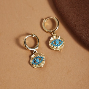 Könnte beinhalten: Ein Paar goldfarbene Creolen mit türkisfarbenem Steindesign. Jeder Ohrring hat einen kleinen Reifen und einen Anhänger mit einem strukturierten Goldrahmen und kleinen türkisfarbenen Steinen. Präsentation vor neutralem Hintergrund.