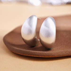 May include: A pair of silver-colored, half-hoop earrings. The earrings have a smooth, rounded design and are displayed on a brown, oval-shaped tray. The earrings have a matte finish and a simple, modern aesthetic.