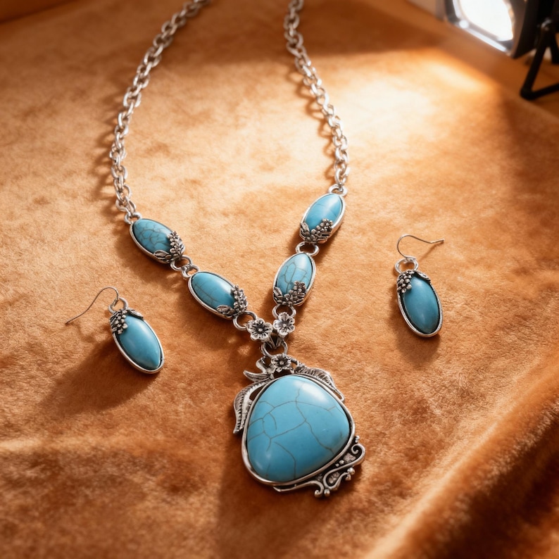 May include: A silver-toned necklace and earring set with turquoise-colored stones. The necklace features a large, teardrop-shaped pendant and smaller oval stones. The earrings have matching oval stones. The jewelry is set against a brown background.