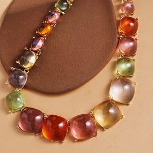 May include: A colorful necklace featuring square-cut gemstones in shades of green, purple, orange, and pink. The gemstones are set in a gold-toned metal chain. The necklace is displayed on a brown surface.