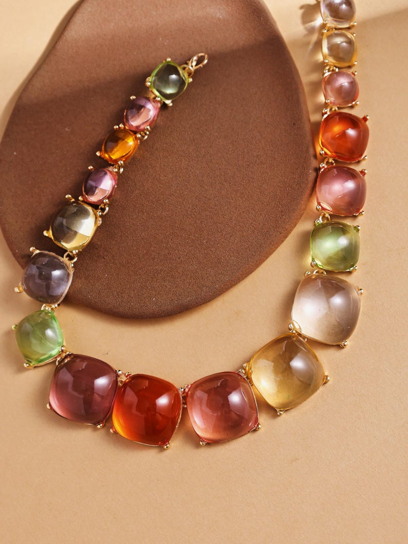 May include: A colorful necklace featuring square-cut gemstones in shades of green, purple, orange, and pink. The gemstones are set in a gold-toned metal chain. The necklace is displayed on a brown surface.