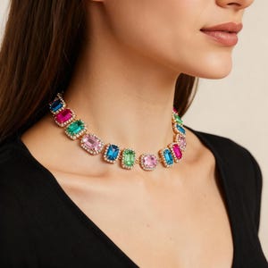 May include: A gold-toned choker necklace featuring a series of colorful, rectangular gemstones. The gemstones are in shades of pink, blue, green, and magenta, each surrounded by a halo of small, clear crystals. The necklace is worn around the neck.