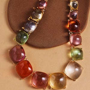 May include: A colorful necklace featuring large, square-shaped, translucent beads in shades of red, orange, yellow, green, and purple. The beads are connected by gold-colored metal links, creating a vibrant and eye-catching accessory.