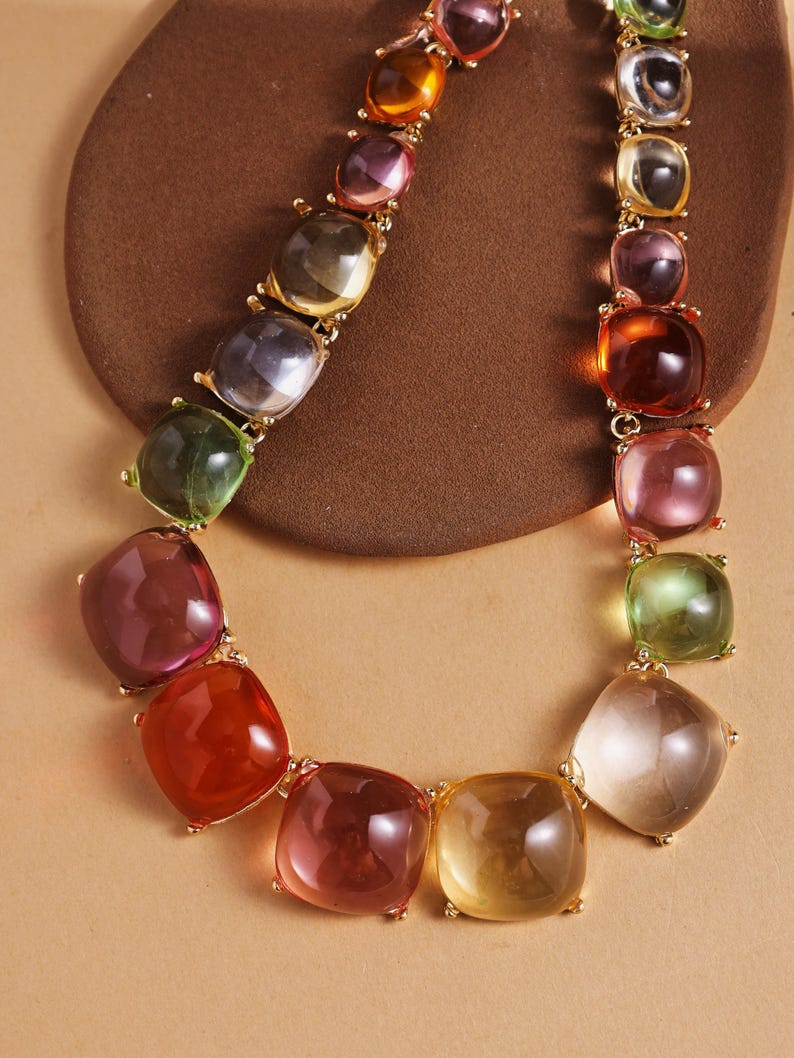 May include: A colorful necklace featuring large, square-shaped, translucent beads in shades of red, orange, yellow, green, and purple. The beads are connected by gold-colored metal links, creating a vibrant and eye-catching accessory.