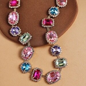 May include: A colorful statement necklace featuring a gold chain and a series of large, multi-colored gemstones. The gemstones are in various shapes, including oval and rectangular, and are surrounded by small, clear crystals. The necklace is displayed on a brown surface.