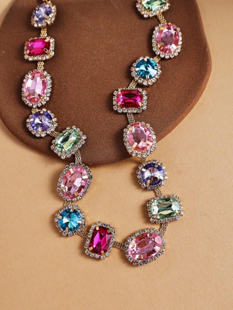 May include: A colorful statement necklace featuring a gold chain and a series of large, multi-colored gemstones. The gemstones are in various shapes, including oval and rectangular, and are surrounded by small, clear crystals. The necklace is displayed on a brown surface.