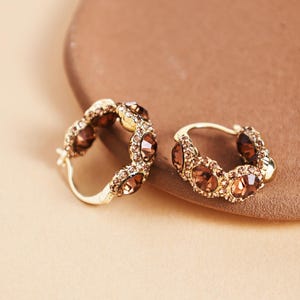 Peut inclure: Une paire de boucles d'oreilles créoles dorées. Chaque boucle d'oreille est ornée d'une série de pierres précieuses ovales de couleur marron. Les boucles d'oreilles sont posées sur un fond neutre, mettant en valeur leur design et leur couleur.