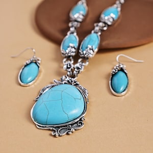 May include: A turquoise jewelry set featuring a necklace and matching earrings. The necklace has a large, teardrop-shaped turquoise pendant with a silver-colored filigree setting. The earrings mirror the pendant's shape. The set is displayed on a neutral background.