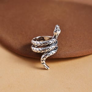 Puede incluir: Un anillo plateado con forma de serpiente enrollada. La cabeza de la serpiente está levantada y el cuerpo se enrolla alrededor del dedo. El anillo tiene escamas detalladas y una superficie texturizada, creando una apariencia realista. El anillo está sobre un fondo marrón.