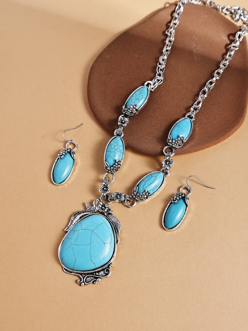 May include: A silver-toned necklace and earring set featuring turquoise-colored stones. The necklace has a large, oval pendant and smaller oval stones along the chain. The earrings are oval-shaped and dangle from hooks. The jewelry has a vintage, bohemian style.